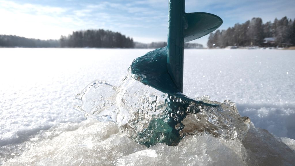 Bilden föreställer en turkos isborr som håller på att dras upp ur hålet man borrat med den. Man befinner sig ute på en sjö, med snötäckt is, och solen lyser. Längs sjökanten syns skog, och även ett hus en bit bort.