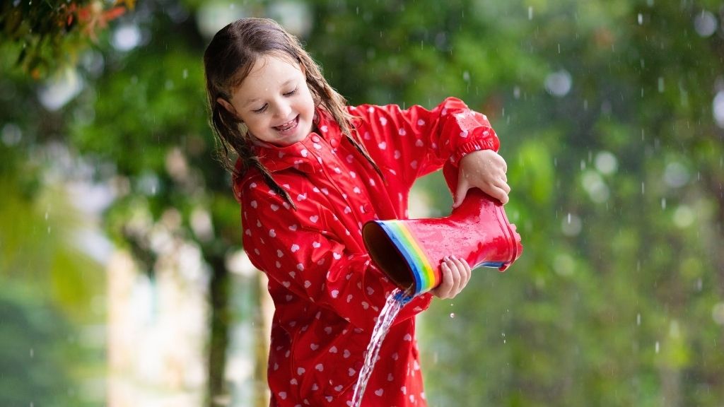 Bilden föreställer en flicka som leker i regnet. Hon har en röd regnjacka med små rosa hjärtan på. Hon håller på att hälla vatten ur en röd stövel med en regnbågsfärgad rand högst upp på skaftet.