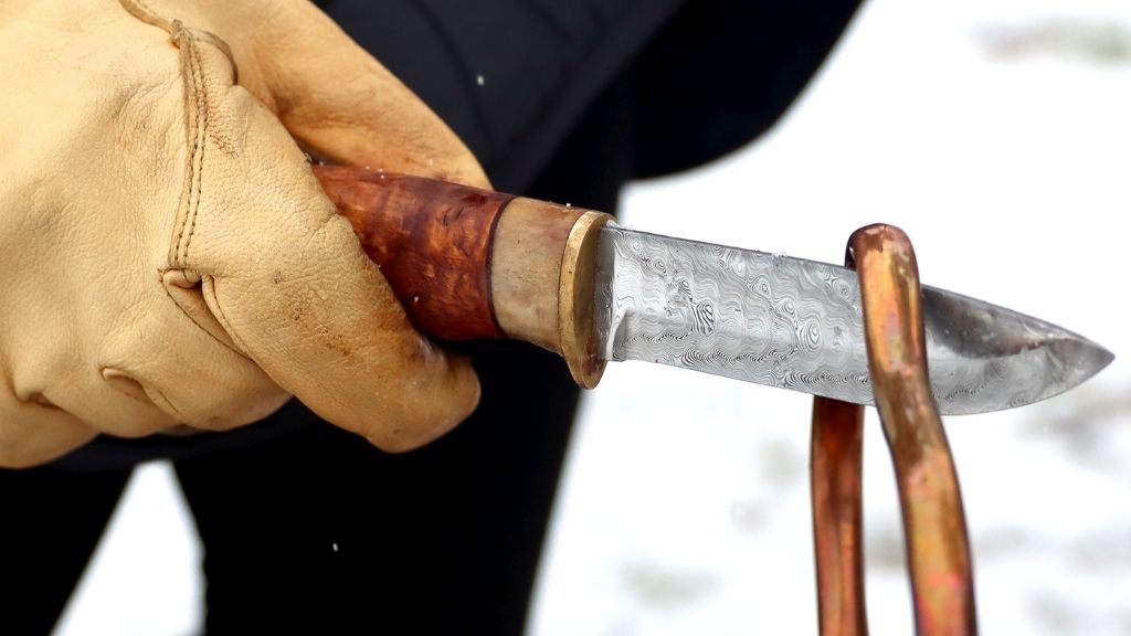 Handskbeklädd hand som håller i en friluftskniv med träskaft som håller handtag till kopparkittel