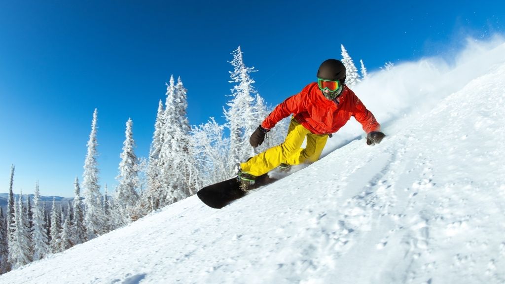 Snowboardåkare i röd jacka och gula byxor i kraftigt lutande backe med blå himmel bakom