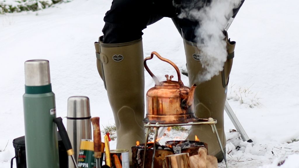 Gröna gummistövlar i vintermiljö bakom en rykande kopparkittel och ett par termosar