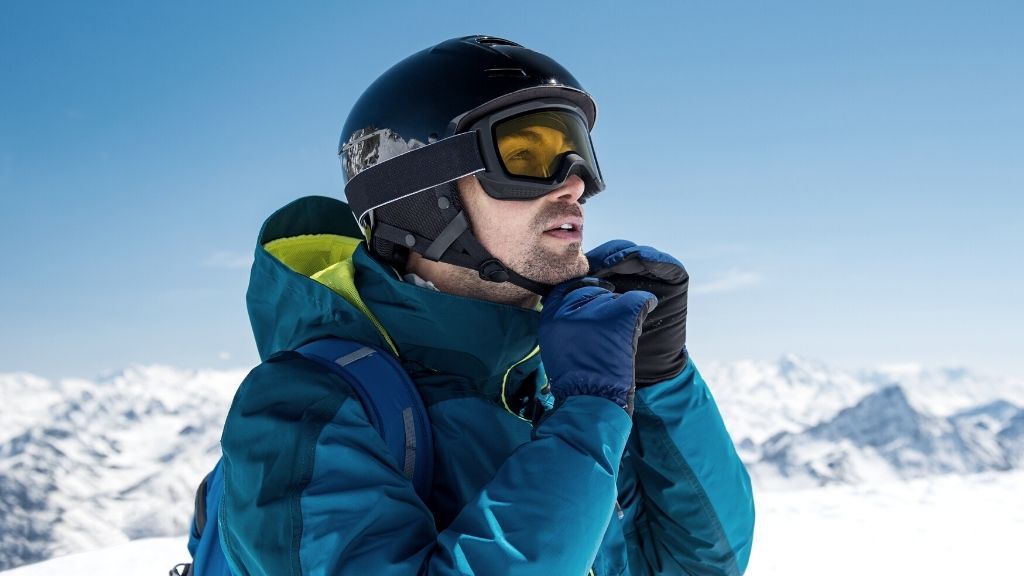 Bilden föreställer en man som befinner sig högt uppe på ett fjäll. Han har skidkläder, ryggsäck, vantar och skidglasögon på sig. Han håller på att knäppa sin skidhjälm.