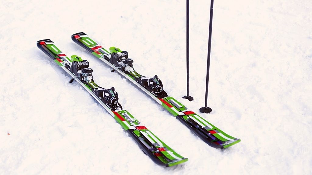 Bilden föreställer ett par skidor som ligger på en snötäckt mark. Bredvid skidorna står ett par skidstavar nedstoppade i snön.