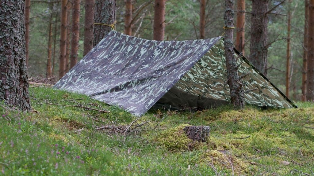 Bilden föreställer ett kamouflage-mönstrat vindskydd, som står uppfällt i en skog.