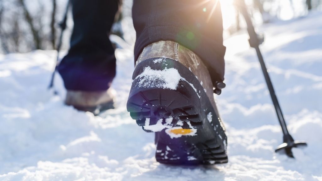 Bilden föreställer en närbild av ett par vinterskor. Personen som bär dem är ute och går i ett vinterlandskap med snö. Personen använder även vandringsstavar.
