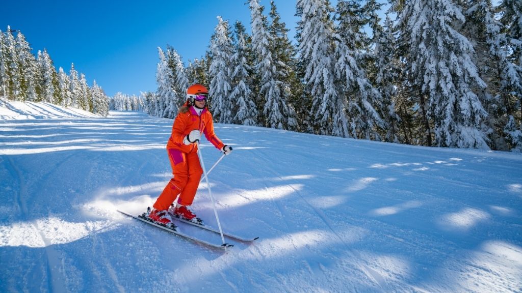 Bilden föreställer en kvinna som åker skidor, iförd en orange skidjacka med rosa detaljer. Hon har också på sig tillhörande byxor i samma färg. Hon bär också en orange hjälm och har skidglasögon vars visir skimrar i rosa. Hon åker ned för en backe som är kantad av snöbetäckta granar.