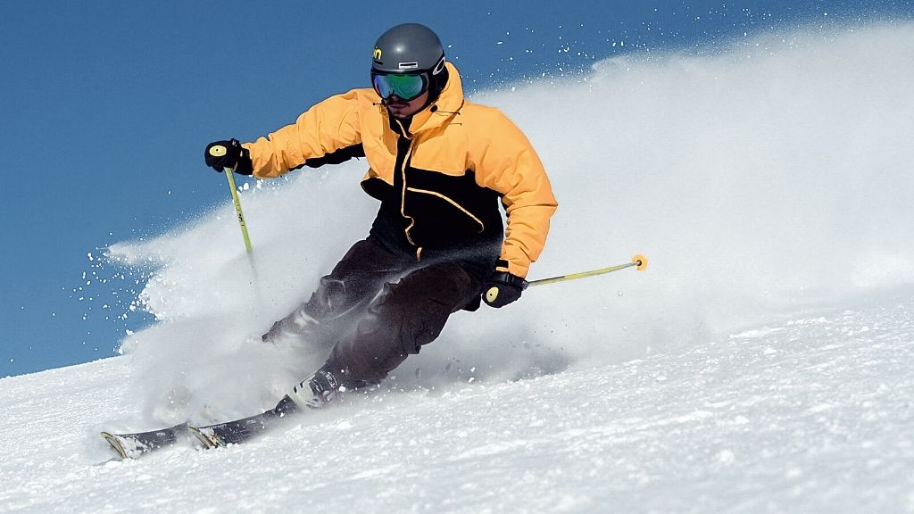 Bilden föreställer en man som åker slalomskidor, och snön yr bakom honom. Han har svarta skidbyxor, och en skidjacka i svart och gult. Han bär även skidglasögon och skidhjälm.