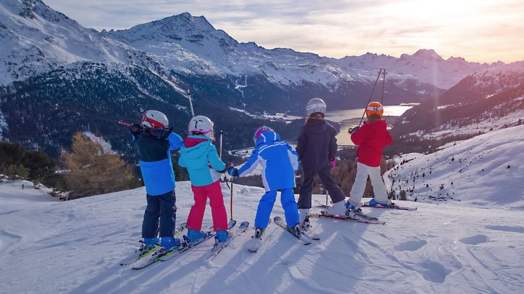 Bilden föreställer fem barn som står uppe på ett fjäll och blickar ner över den underliggande dalen. Alla barnen bär skidor och har skidkläder på sig. Det verkar vara senare på dagen, då solen håller på att gå ned.